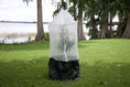 Load image into Gallery viewer, Skirted Tree Defender protecting a young tree on a grassy lawn beside a lake. The breathable mesh cover includes a front zipper and a black skirt secured around the base.
